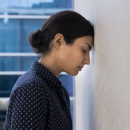 A young person with their hair in a bun leaning their forehead against a white wall in a business environment.