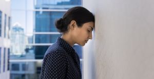 A young person with their hair in a bun leaning their forehead against a white wall in a business environment.