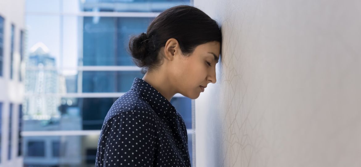 A young person with their hair in a bun leaning their forehead against a white wall in a business environment.