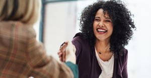 A smiling woman wearing business casual attire reaches forward to shake the hand of a person across the table.