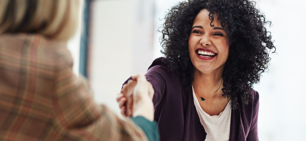 A smiling woman wearing business casual attire reaches forward to shake the hand of a person across the table.
