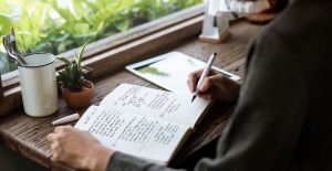 A person sitting at a wooden desk and reviewing notes in a small journal. They hold a pen just above one of the pages.