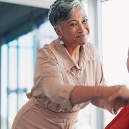 A businesswoman in a muted pink jumpsuit reaches forward to shake hands with another woman in a the workplace.
