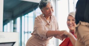 A businesswoman in a muted pink jumpsuit reaches forward to shake hands with another woman in a the workplace.