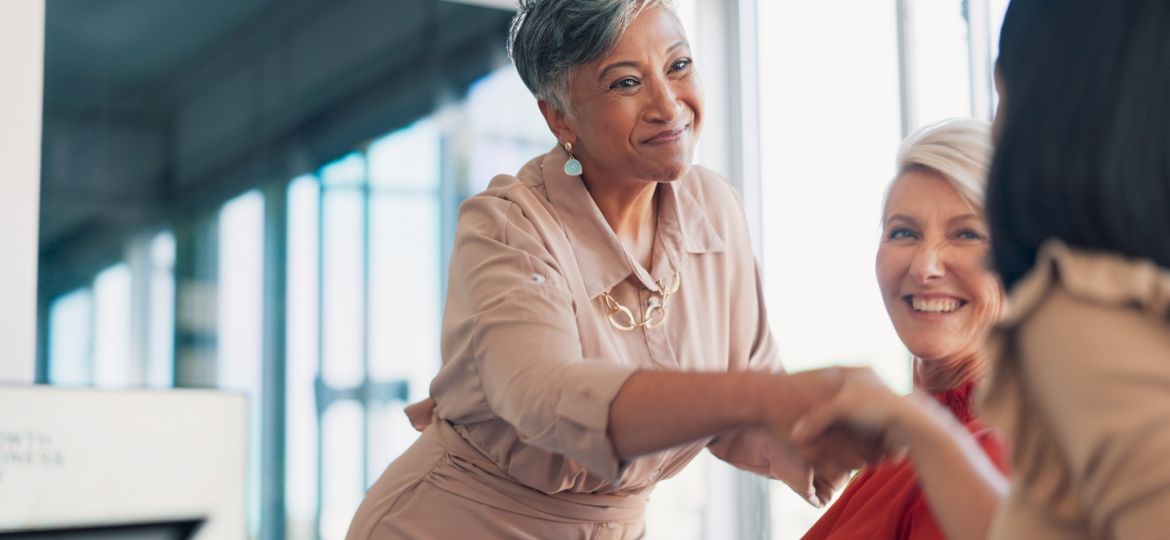 A businesswoman in a muted pink jumpsuit reaches forward to shake hands with another woman in a the workplace.