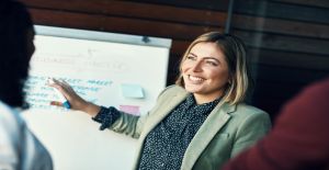 A woman stands in front of a whiteboard covered in writing and sticky notes. She grins as she talks to her employees.