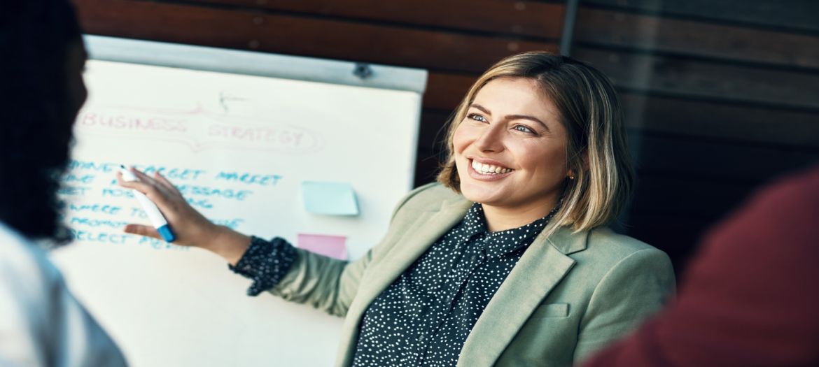 A woman stands in front of a whiteboard covered in writing and sticky notes. She grins as she talks to her employees.