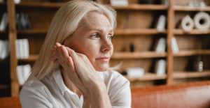 An aging woman sits on a leather sofa as she stares into the distance. She has a stressed look on her face.