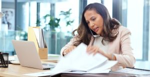 A businesswoman looks frazzled as she holds the phone up to her ear and shuffles through papers on her desk.