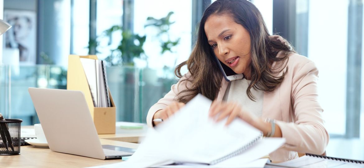 A businesswoman looks frazzled as she holds the phone up to her ear and shuffles through papers on her desk.