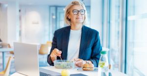 A businesswoman smiles as she sits at a table in an employee lounge and eats a salad out of a container.