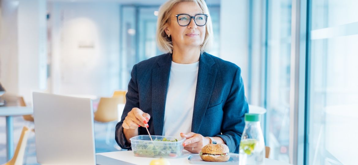 A businesswoman smiles as she sits at a table in an employee lounge and eats a salad out of a container.