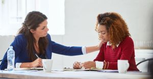 A businesswoman consoles a co-worker who looks very sad. The two women sit at a table, with papers and cups.