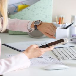 A professional woman checking her wristwatch while she sits at her office desk and holds her smartphone.