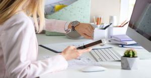A professional woman checking her wristwatch while she sits at her office desk and holds her smartphone.