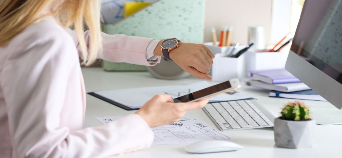 A professional woman checking her wristwatch while she sits at her office desk and holds her smartphone.