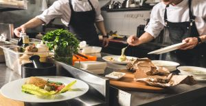 A close-up of two chefs wearing black aprons, preparing three meals on white plates inside a large kitchen.