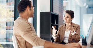 A business manager talks to her employee as they sit in her office. She has her hands up while he smiles and listens.