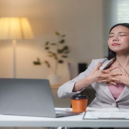 A young business woman sits at a desk in an office. She closes her eyes, interlocks her fingers, and takes deep breaths.