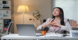 A young business woman sits at a desk in an office. She closes her eyes, interlocks her fingers, and takes deep breaths.