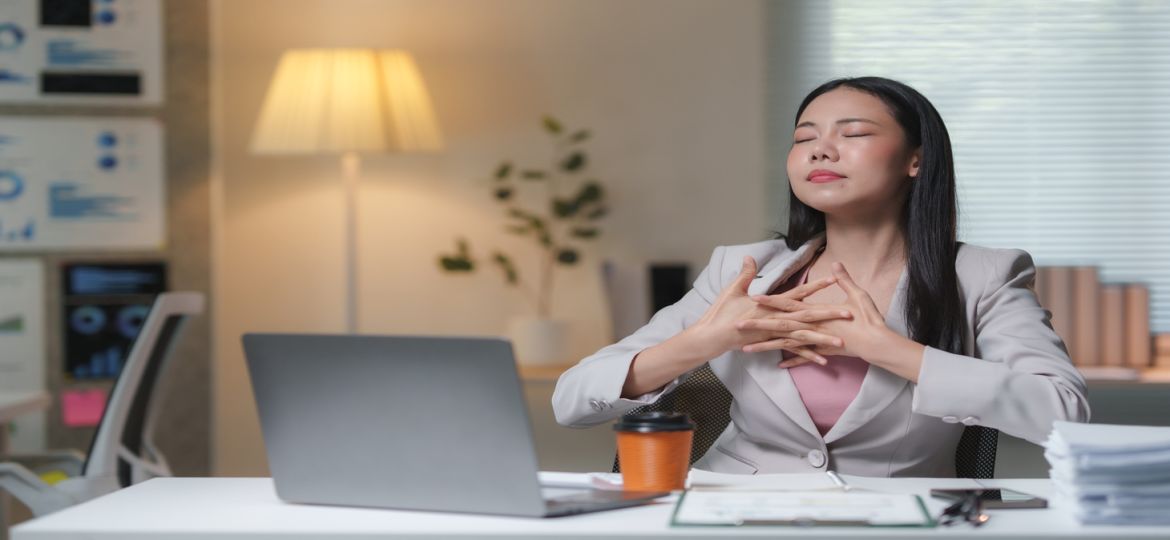 A young business woman sits at a desk in an office. She closes her eyes, interlocks her fingers, and takes deep breaths.