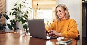 A woman wearing wire-frame glasses and a yellow sweater smiles in front of a laptop with a phone and notebook sitting next to it.