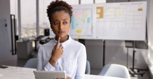A young businesswoman stands in an office, thinking about something with her finger on her chin. She holds a tablet.