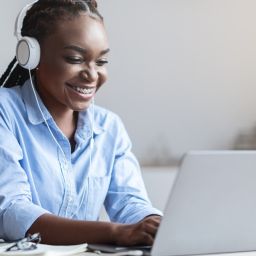 A woman smiles as she wears headphones and works on her laptop at her desk. A gray mug sits on the desk.