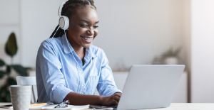 A woman smiles as she wears headphones and works on her laptop at her desk. A gray mug sits on the desk.