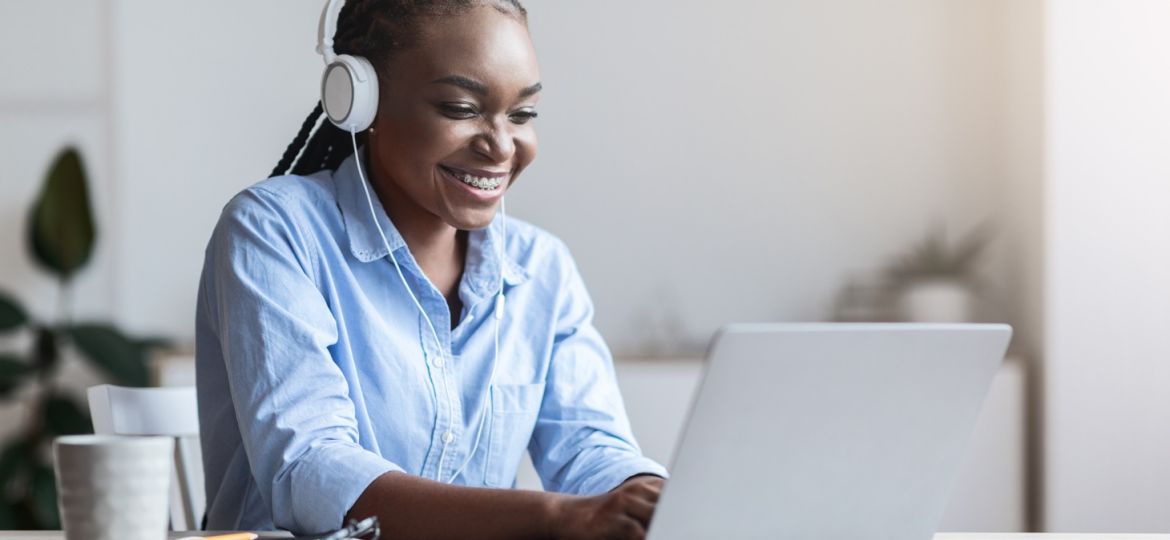 A woman smiles as she wears headphones and works on her laptop at her desk. A gray mug sits on the desk.